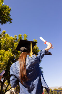Graduating student is raising diploma scroll while wearing mortarboard against bright blue sky