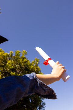 Arm is holding rolled diploma tied with red ribbon, rising past gown sleeve near blue sky