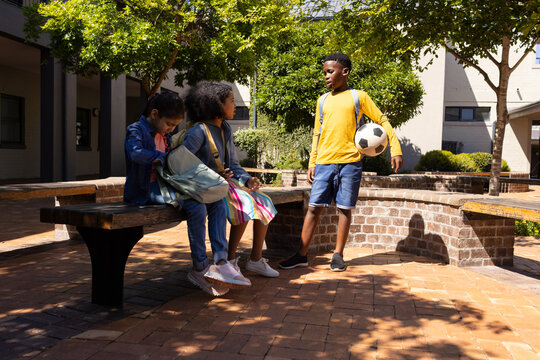 Diverse youth friends chatting on campus courtyard by wooden bench with soccer ball and backpacks