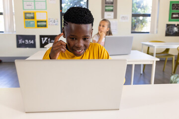 Diverse teenage students sitting at desks in classroom, male in yellow shirt using silver laptop