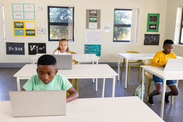 Diverse students working solo at classroom desks using laptops near yellow chairs and math posters