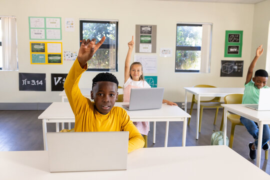 Diverse teenage classmates raising hands in classroom at white desks using laptops