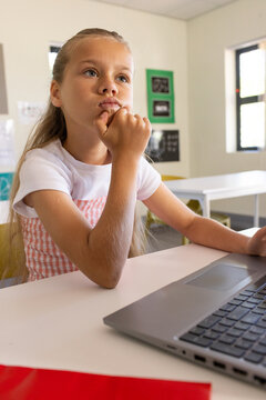 Preteen female student sitting at classroom table, wearing pink dress, gazing near silver laptop