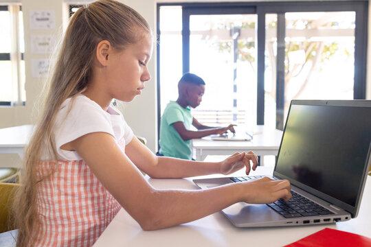Diverse classmates sitting and typing on silver laptop and using device in class near red notebook
