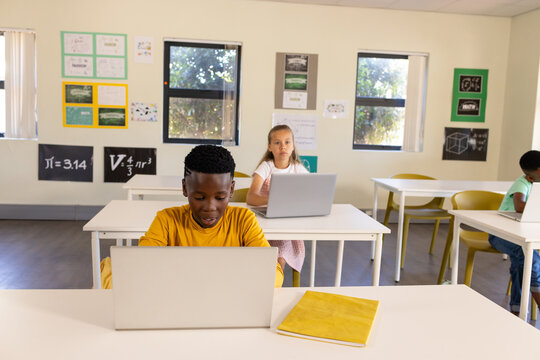 Diverse classmates working on silver laptops in class with mustard shirt using yellow notebook