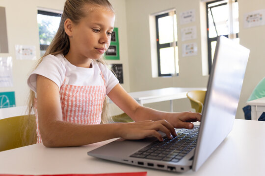 Preteen girl wearing pink pinafore sitting at classroom desk using laptop, red folder nearby
