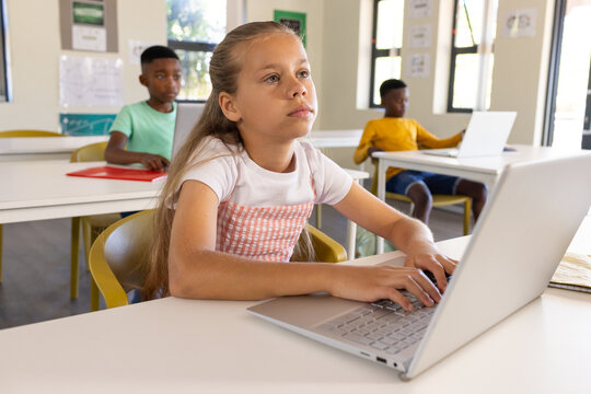 Diverse youth students sitting in classroom, female youth in pink dress typing on silver laptop