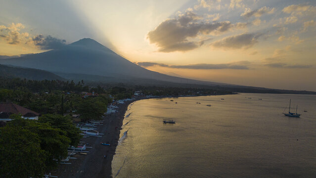 Aerial view of the dark, volcanic sand beach kissed by gentle waves, shadowed by the majestic Mount Agung under a golden sunset, Amed, Bali, Indonesia.