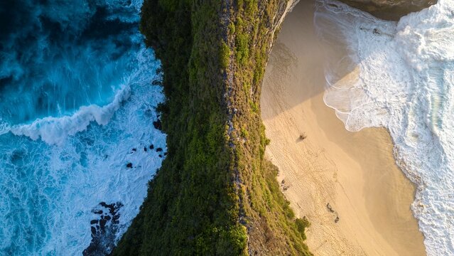 Aerial view of the craggy, verdant cliffs meet the turquoise sea and soft, sandy beach at Nusa Penida Island, Bali, Indonesia.