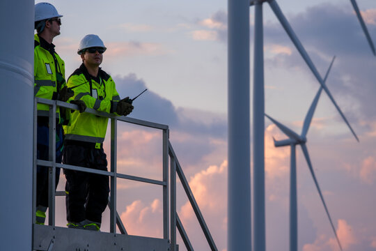 Two wind turbine engineers inspect farm windmills during early evening in the United States