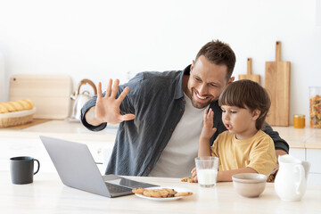 Online communication. Little boy and his daddy video calling to relatives via laptop, waving hands to computer and smiling, sitting together at kitchen, free space