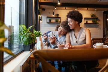 African-American woman and her son sit in cafe and use smartphone, teenager plays on mobile phone and smiles with his mother in restaurant