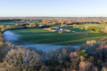 Aerial view of a verdant field embraced by a frost-kissed rim and a backdrop of dense woodland under a serene sky, Yateley, England, United Kingdom.