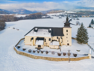 Aerial view of a quaint, cream-colored church standing resilient against a pristine white snow-covered landscape, framed by distant mountains, Lopej, Banskobystricky kraj, Slovakia.