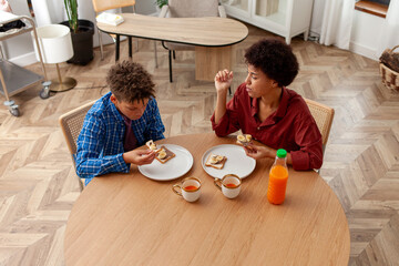 African American woman eating chocolate butter banana sandwiches and juice with her son at home at the table, teenager having lunch with his mother and laughing
