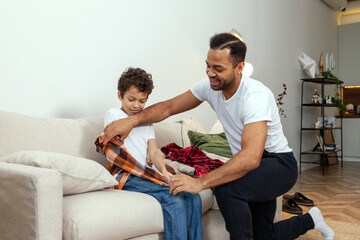 African American man helps his son put on his shirt in the morning, father takes care of the child and gets him ready for school