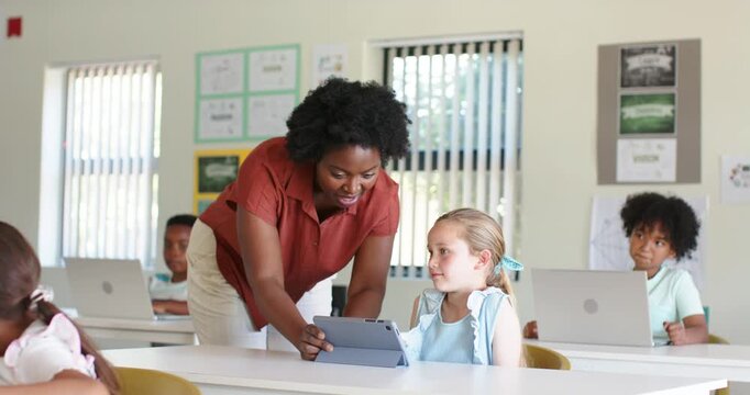 African American woman teacher noticing youth girl needing help, leaning over tablet in classroom