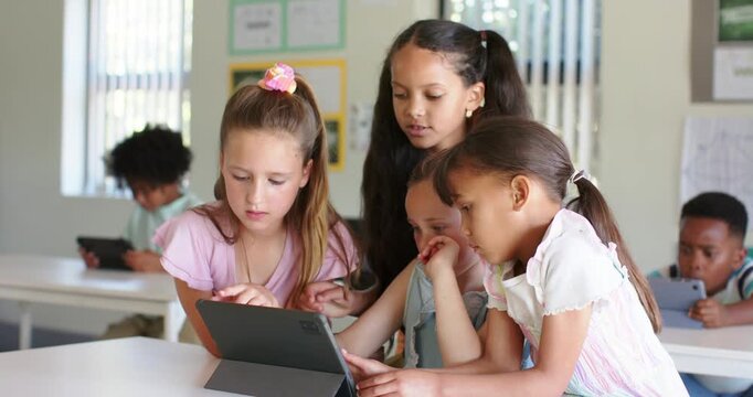 Four school-age girls gathering, left girl tapping tablet, collaborating on math task in classroom