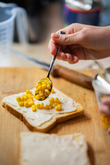 Hand Spooning Sweet Corn onto White Bread with Creamy Spread on a Wooden Board