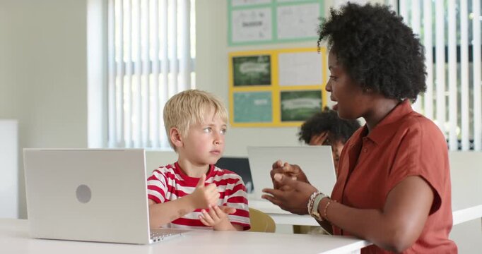 African American woman raising thumb, teaching boy finger counting at school with silver laptop