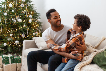 African American man and his son hug on the couch at home on New Year's Eve, teenager talks to his father and smiles against the backdrop of Christmas tree
