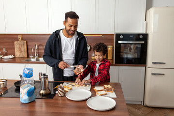 African American man and his son prepare sweet sandwiches with chocolate and banana in the kitchen, father and child have breakfast together at home