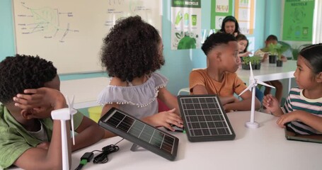Diverse kids watching girl moving solar panel in STEM class with turbine and tablet teacher nearing