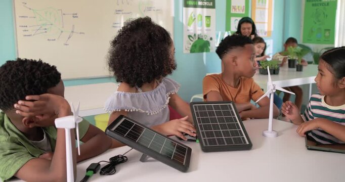 Diverse kids watching girl moving solar panel in STEM class with turbine and tablet teacher nearing