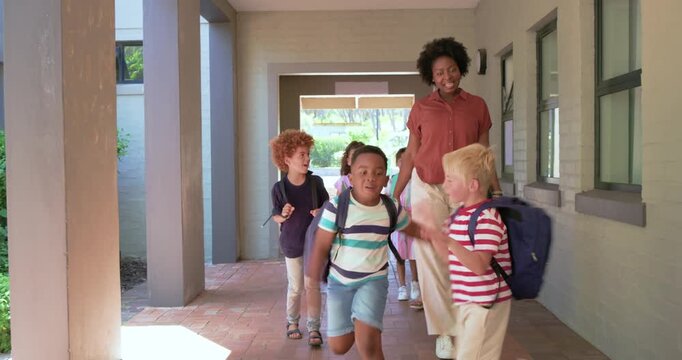 African American teacher guiding kids to sunlight through covered hall, holding tablet, backpacks