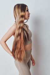 Naklejka premium Young woman with long straight hair standing sideways wearing beige sportswear indoors. Female model with casual pose against plain background showing natural hairstyle and fitness outfit.