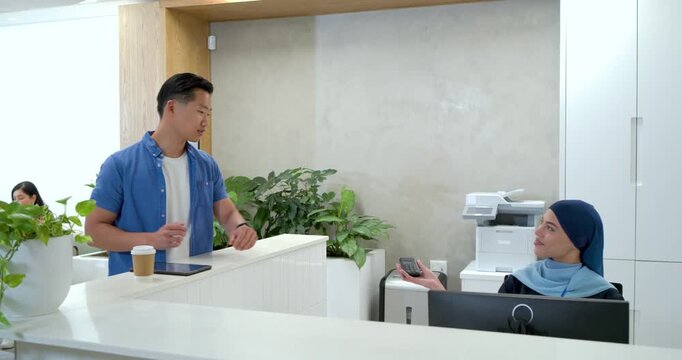 Asian man 30s tablet or coffee checking watch extending card at desk to receptionist hijab checking 