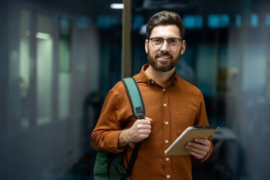 Portrait of a young man wearing glasses and carrying a backpack standing indoors, holding a notebook in his hand and looking at the camera with a smile
