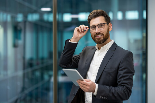 Portrait of a young successful businessman in a suit and glasses standing in the office, holding a tablet in his hand and looking at the camera with a smile