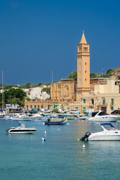 The Parish Church of St. Anne with view of Marsaskala Harbour with blue sky and turquoise water. Malta.