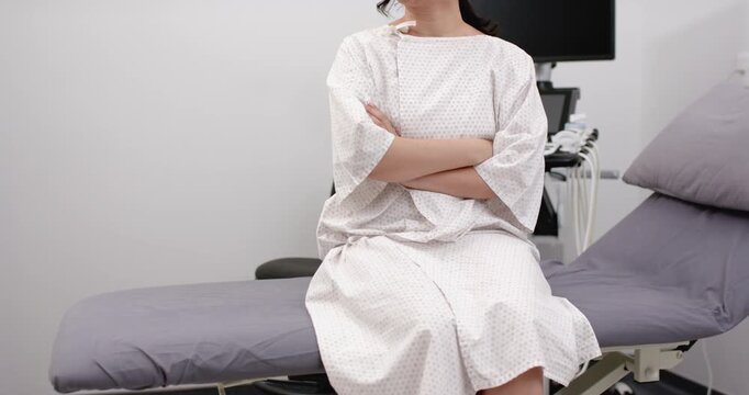 Woman sitting on exam bench in clinic, camera framing shifting, adjusting gown, waiting for exam