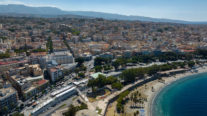 Fototapeta premium Aerial view of the city of Reggio Calabria, Southern Italy. It is a coastal Mediterranean town. Densely packed buildings with red-tiled roofs meet a curved sandy beach and deep blue sea.