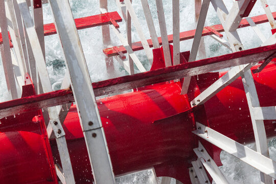 Centered view of a riverboat paddlewheel hub painted red, framed by intersecting silver struts and splashing water for a dynamic industrial composition.