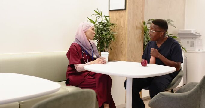 Med staff in scrubs sitting at lounge, chatting on break, holding cup and apple, checking wrist