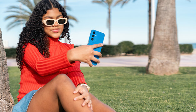 Young woman taking selfie with smartphone while sitting on grass in a park near the beach