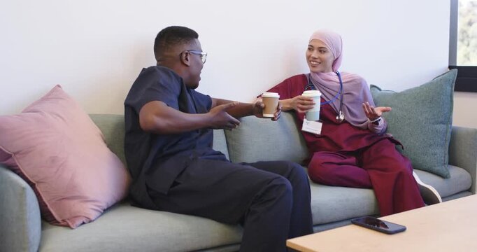 Diverse coworkers on break in scrubs, chatting then laughing on staff lounge couch with coffee cups