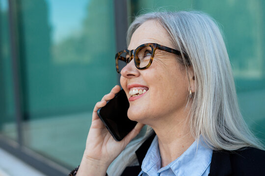 Smiling businesswoman talking on smartphone standing outdoors near modern office building
