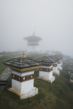 Aerial view of the serene Dochula Chorten shrouded in mist, its white stupas and dark roofs contrasting against the soft, diffused light, Dochula, Thimphu, Bhutan.