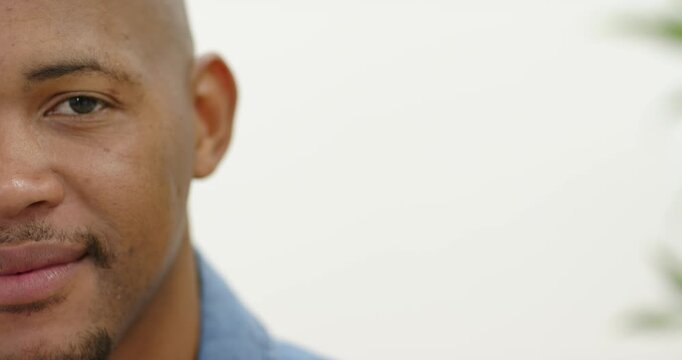 Man holding eye contact, shifting to smile while wearing blue collared shirt in room near plant