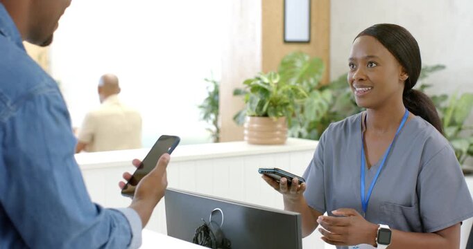 African American woman at clinic desk readying scanner as visitor extending phone, handling payment
