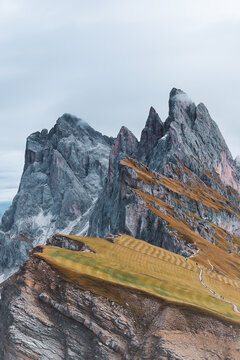 Aerial view of jagged peaks rise dramatically against a muted sky, their rocky faces contrasting with the golden hues of the lower slopes, Seceda, Italy.