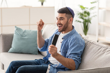 Portrait of excited young guy watching football match, shaking clenched fists. Happy jewish man...