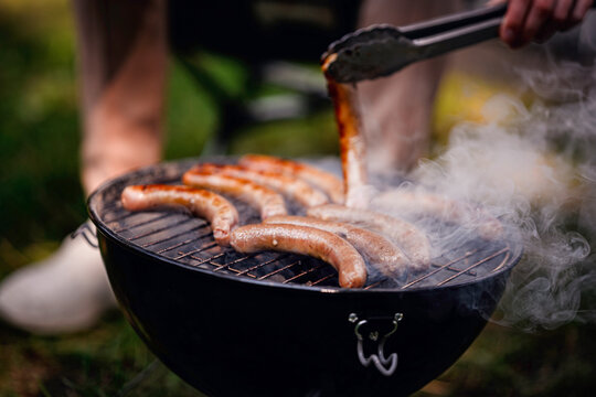 Close-up of hand using tongs to flip sizzling sausages on a smoky charcoal grill during outdoor cooking.