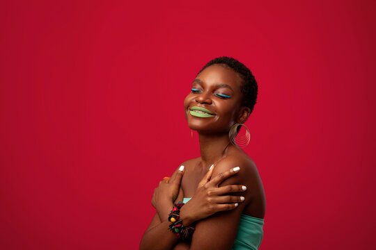 Exotic afro american young woman posing on red studio background, black female model wearing bright outfit and funky makep, using stylish accessories, hugging her shoulders and smiling, copy space