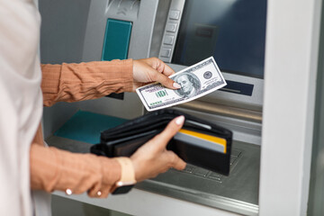 Woman using modern ATM, taking money from cash machine outdoors, closeup. Covid-19 pandemic. Young unrecognizable arabian woman holding dollars and wallet with credit cards on street, free space