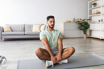 Peaceful millennial Arab guy sitting in lotus pose, meditating with closed eyes on mat at home,...